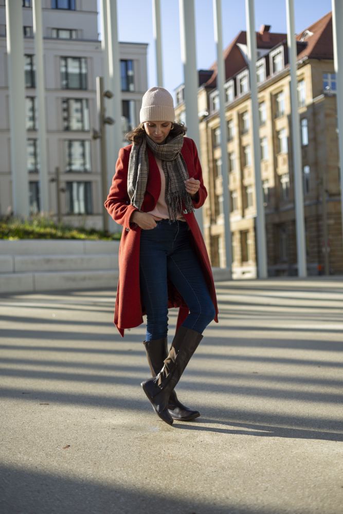 Eine Person in rotem Mantel und Schal geht eine sonnige Stadtstraße entlang. Sie trägt Jeans und Stiefel in Nappabraun von ZAQQ sowie eine Beanie. Hohe Gebäude werfen Schatten und verleihen der lebhaften Szene Tiefe.