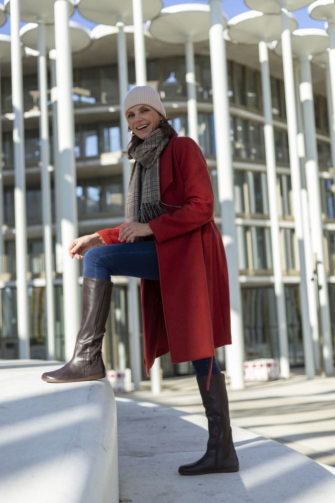 Eine Person in rotem Mantel, Schal und Beanie posiert im Freien mit einem Fuß auf einer ZAQQ QATCH Nappa Brown-Oberfläche. Sie lächelt, trägt Jeans und hohe Damenstiefel. Im Hintergrund sind ein modernes Glasgebäude und ein klarer blauer Himmel zu sehen.