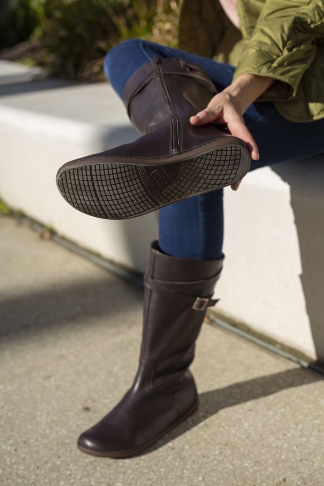 Eine Person sitzt auf einer Bank. Sie trägt Blue Jeans und die Stiefel ATTRAQT Nappa Brown von ZAQQ. Sie hält ein Bein hoch, sodass die Sohle des Stiefels zu sehen ist, während die Sonne verspielte Schatten auf den Boden wirft.