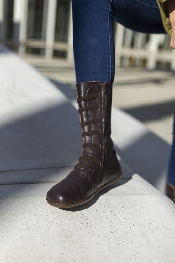 Eine Person in Blue Jeans und dunkelbraunen ATTRAQT Stretch Nappa Kniestiefeln von ZAQQ steht im Sonnenlicht auf einer Betonfläche. Die Stiefel mit mehreren Riemen auf der Vorderseite verleihen einen Hauch von Eleganz. Im Hintergrund sind Schatten und eine Gebäudestruktur zu sehen.