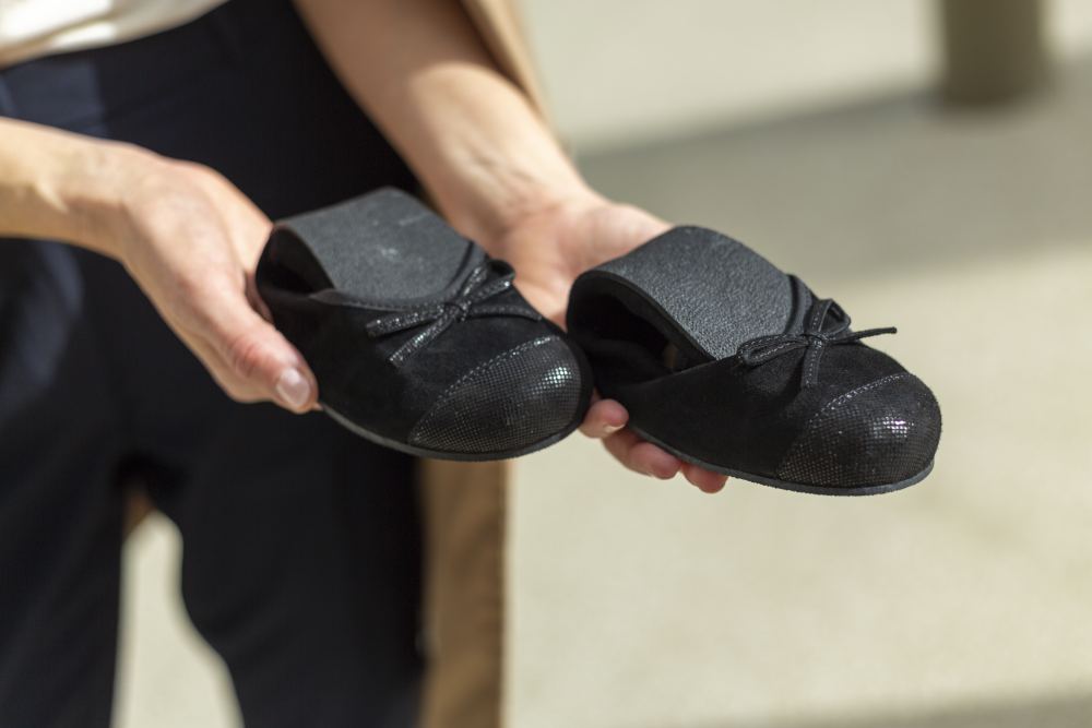 Eine Person hält ein Paar der Ballerinas TWIST Velours Black von ZAQQ mit eleganten Schleifen an der Spitze und präsentiert die Schuhe mit ihrem strukturierten Material aus Veloursleder in einer gut ausgeleuchteten Umgebung dezent in Richtung Kamera.