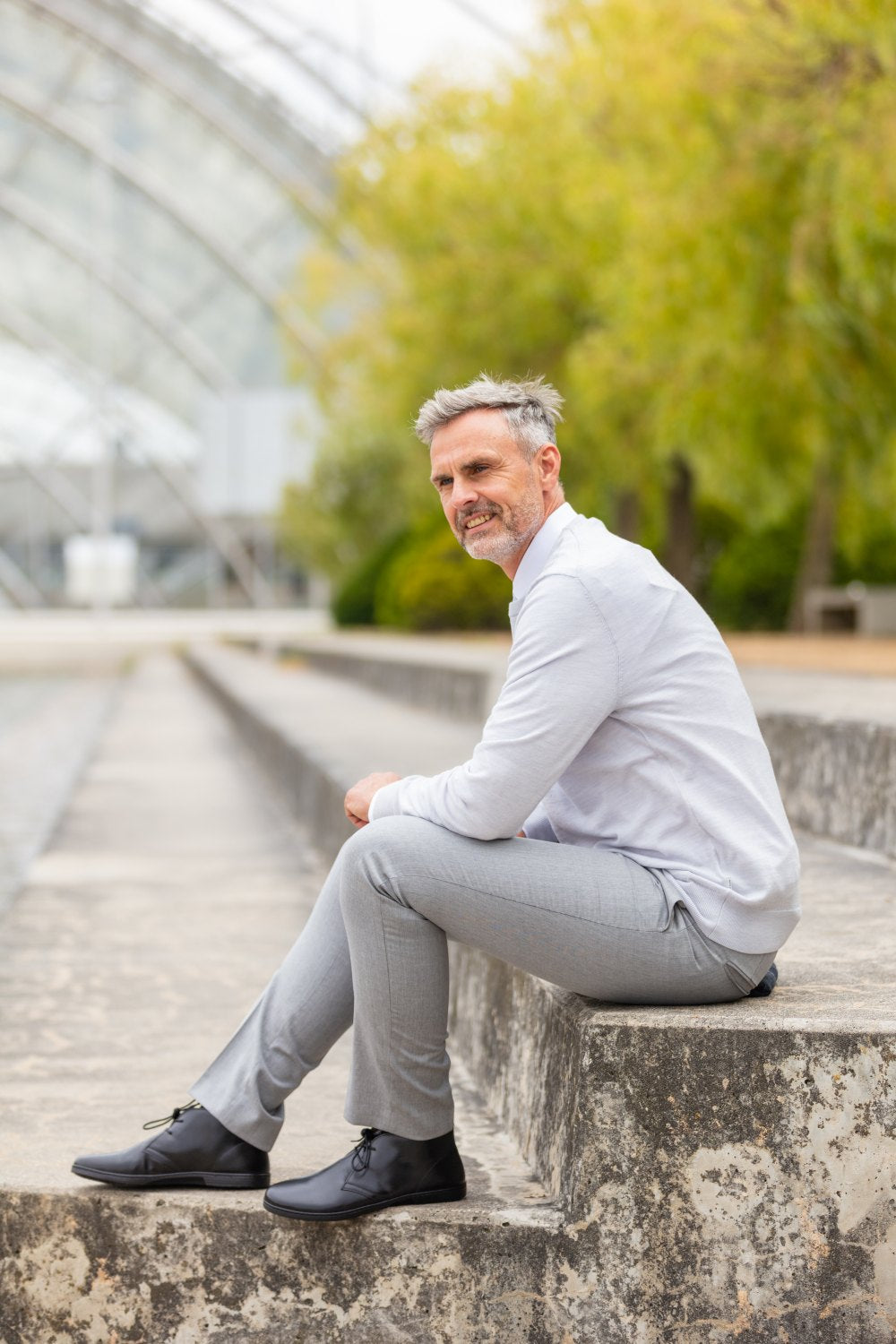 Ein älterer Herr mit grauem Haar, gekleidet in ein weißes Hemd und graue Hosen, sitzt draußen auf Steinstufen und trägt die stylischen schwarzen DUQE-Stiefel von ZAQQ. Er hat ein subtiles Lächeln, während Grünpflanzen und eine Glaskonstruktion die Szene einrahmen.