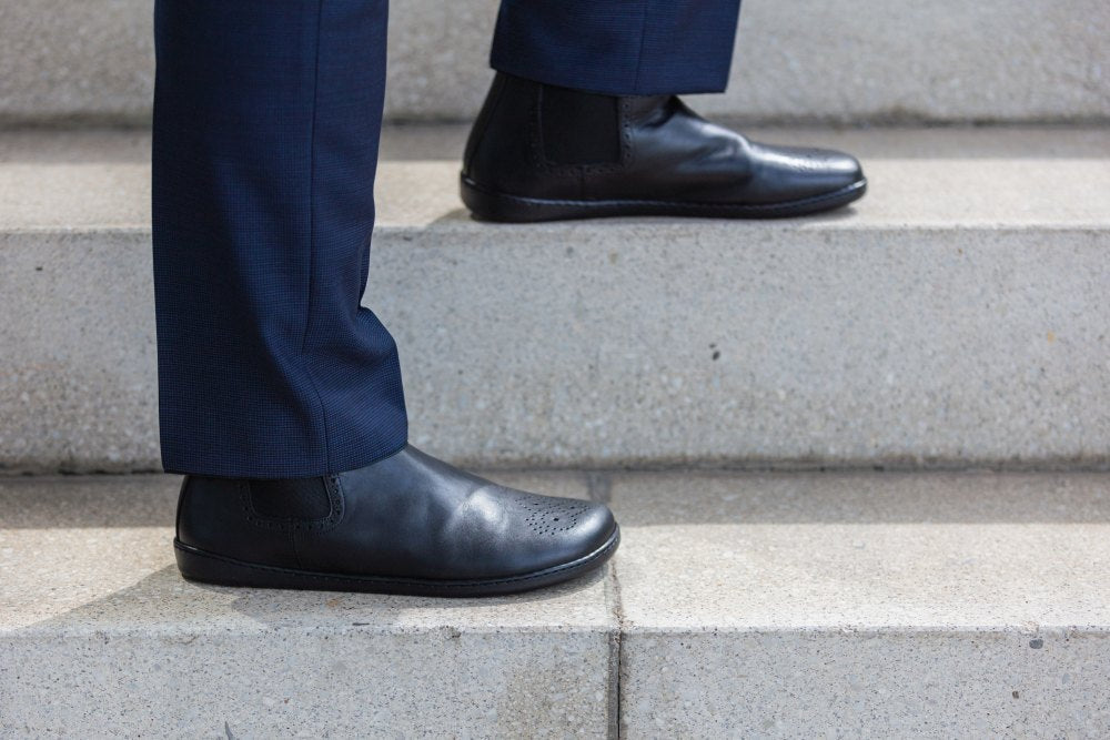 Eine Person in hochwertigen Chelsea-Boots und marineblauer Hose steigt eine Betontreppe hinauf und präsentiert das polierte schwarze Lederschuhwerk und die Unterschenkel. Die Eleganz des ZAQQ EQUITY Brogue Black-Designs wird hervorgehoben.