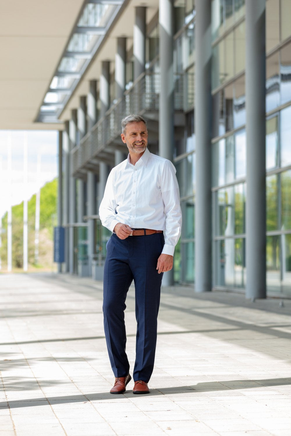 Ein Mann in weißem Hemd und blauer Hose läuft selbstbewusst, in ZAQQ PEAQ Cognac Schuhen mit Barfußsohle, vor einem modernen Gebäude mit großen Fenstern und Säulen. Die Sonne scheint und wirft Licht auf den mit rechteckigen Fliesen gemusterten Weg.