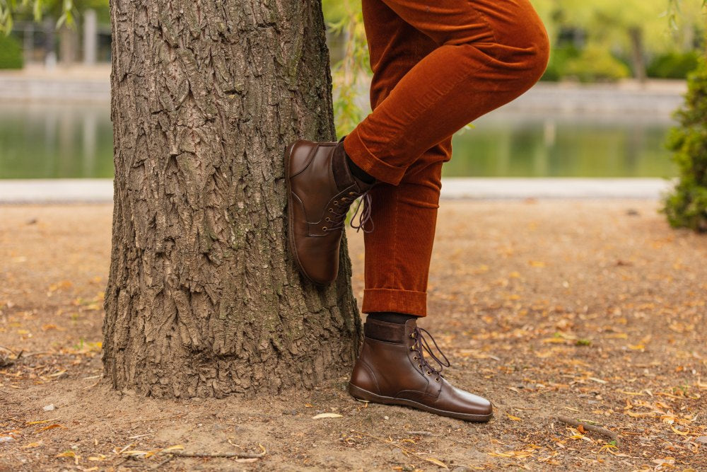 Eine Person in QORBY Brown Boots von ZAQQ und rostfarbener Hose lehnt an einem Baum. Im Hintergrund sind ein See und grüne Blätter zu sehen, was an eine Parklandschaft erinnert. Die Szene vermittelt eine entspannte, herbstliche Atmosphäre.