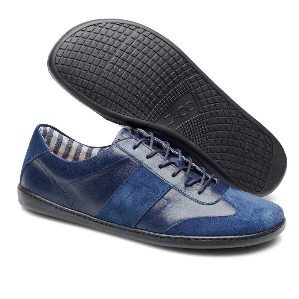 Wir präsentieren die TORQUAY Blue Sneaker von ZAQQ: ein stylisches Paar mit Elementen aus glattem blauem Leder und Velourleder. Ein Schuh steht aufrecht und zeigt seine Seitenteile und Schnürsenkel, während der andere umgedreht ist und eine strukturierte schwarze Sohle mit dem Aufdruck „Zoom“ zeigt. Die Innenseite weist ein Streifenmuster auf, das das Premium-Design unterstreicht.