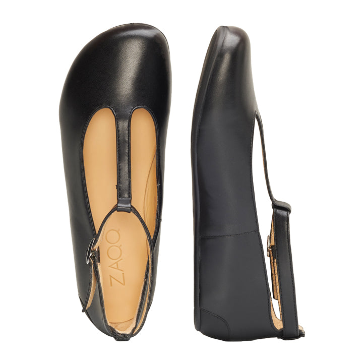 Die QITE Black Flats von ZAQQ mit ihrem raffinierten T-Riemen-Design werden elegant präsentiert. Ein Schuh wird im Seitenprofil und der andere von oben gezeigt, wodurch das beigefarbene Innere und das Zaoq-Branding hervorgehoben werden. Mit einem ergonomisch geformten Fußbett sorgen diese Schuhe bei jedem Schritt für Komfort und Stil.