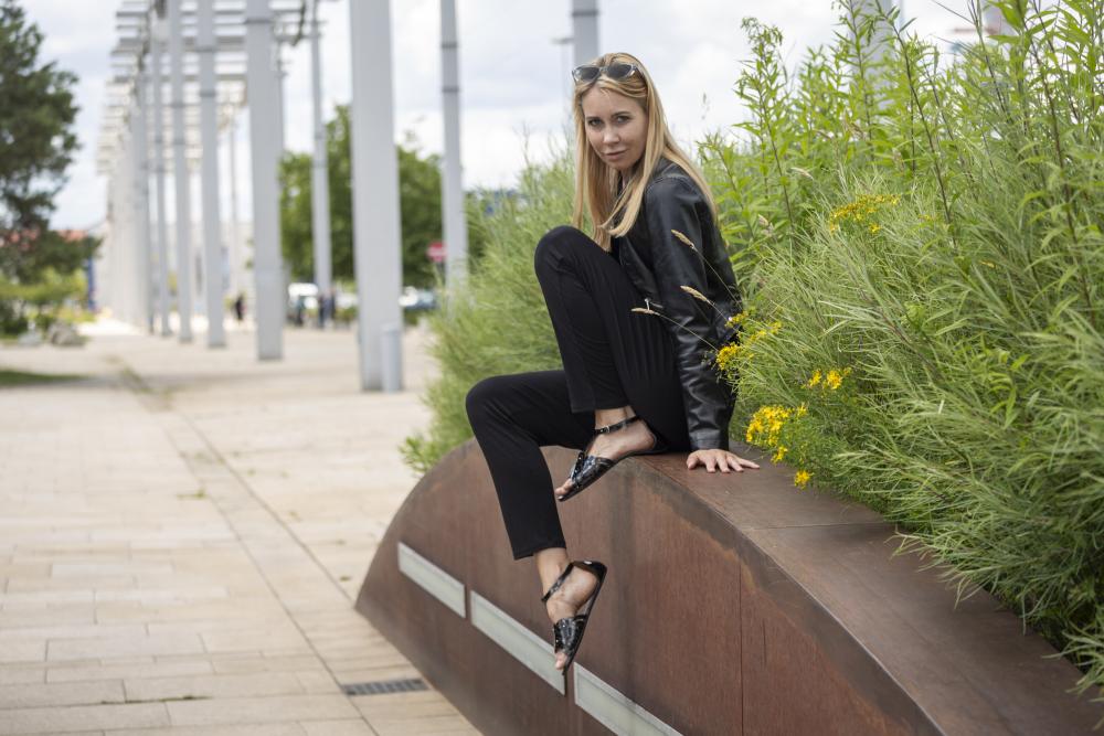 Eine Frau mit langen Haaren, in einem schwarzen Outfit und mit Sonnenbrille auf dem Kopf posiert selbstbewusst auf einer geschwungenen Metallkonstruktion, umgeben von üppigem Grün und gelben Blumen. Ihr schicker Look wird durch die stylischen BOUQET Black Sandalen von ZAQQ komplettiert. Im Hintergrund sind ein gepflasterter Weg und Pfosten zu sehen.