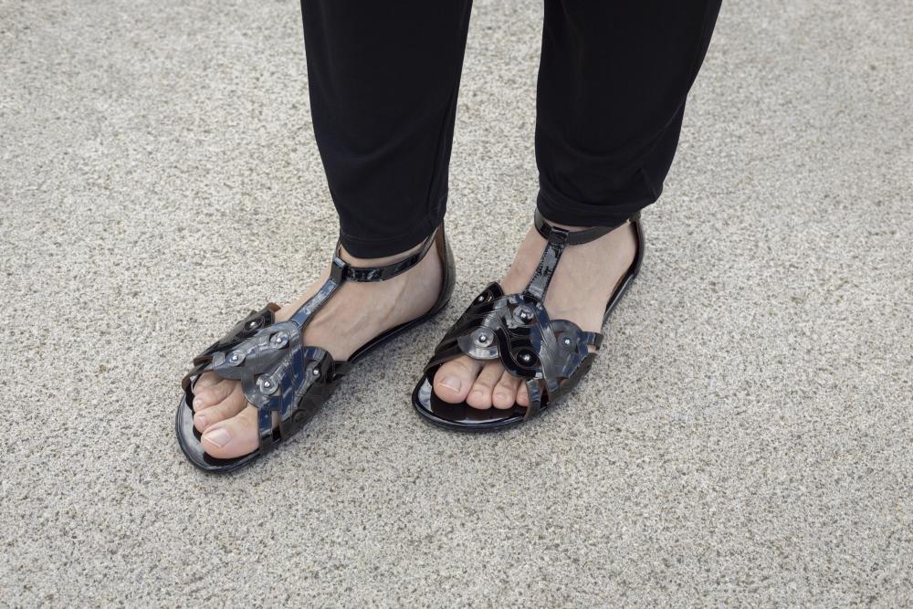 Eine Person in schwarzen Hosen und eleganten BOUQET Black-Sandalen mit Metalldetails von ZAQQ steht selbstbewusst auf einer strukturierten grauen Oberfläche und zeigt die beeindruckende Rutschfestigkeit ihrer Barfußsandale.