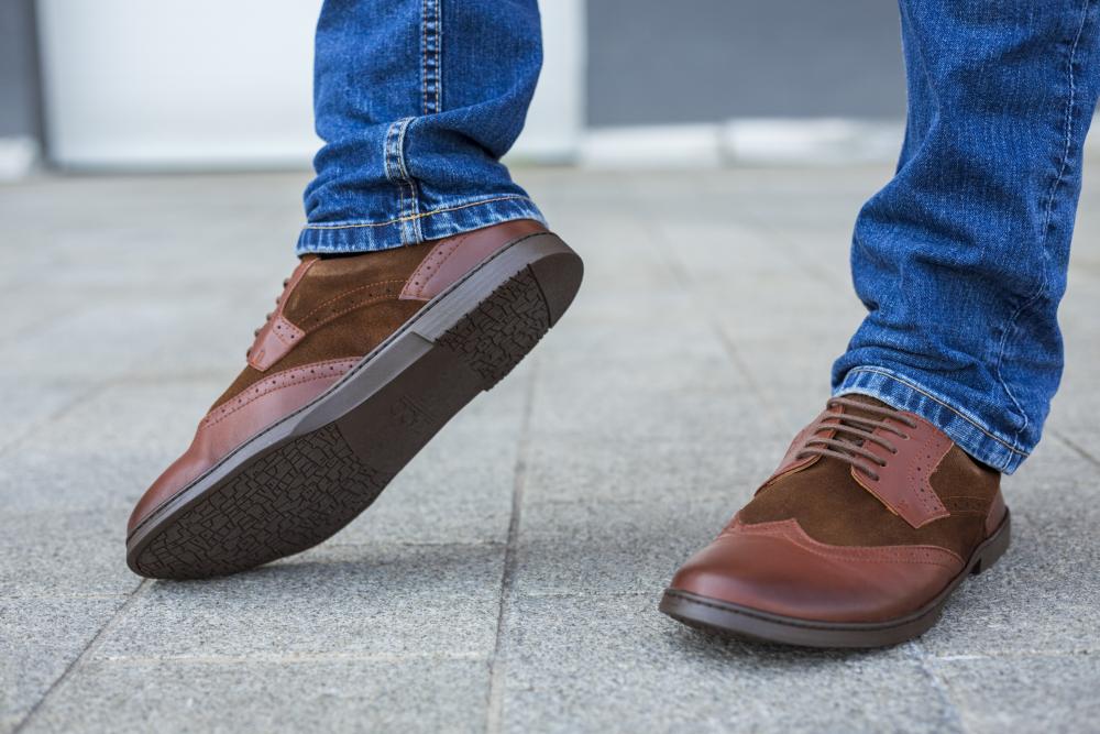 Eine Nahaufnahme zeigt die Füße einer Person, die mit den stilvollen BRIQ Brogue Antique Cognac-Schuhen von ZAQQ geschmückt ist, die aus luxuriösem Kalbsleder und Wildleder gefertigt sind. Die Person steht selbstbewusst auf einer gepflasterten Fläche und kombiniert diese exquisiten Schuhe mit Blue Jeans, um ihren tadellosen Geschmack hervorzuheben.