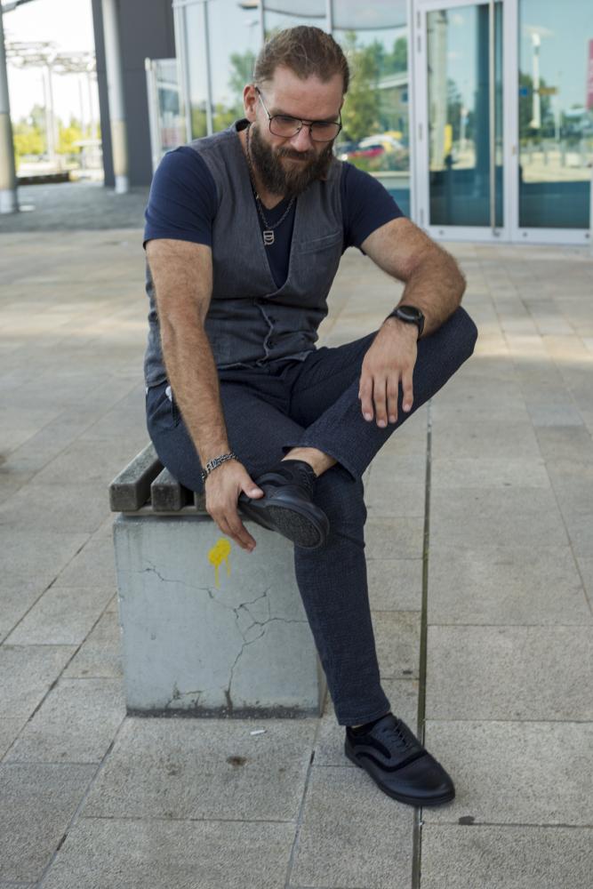 Ein bärtiger Mann mit Brille, dunkler Weste und marineblauem Hemd sitzt draußen auf einem Betonblock und rückt seine BRIQ Brogue Deep Black-Schuhe von ZAQQ zurecht. Im Hintergrund sind ein elegantes Glasgebäude und ein gepflasterter Gehweg zu sehen.