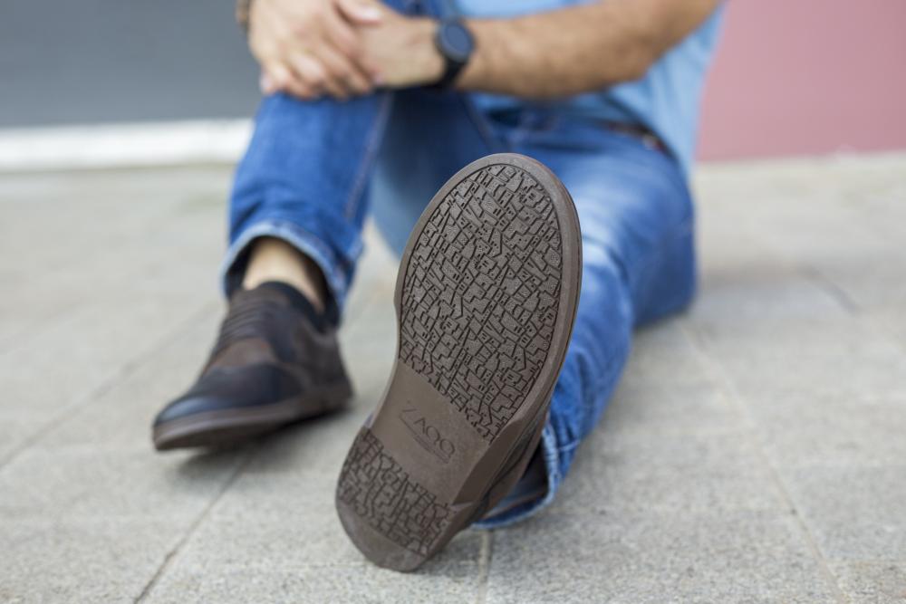 Eine Person sitzt mit gekreuzten Beinen auf dem Boden und trägt Jeans und ein Paar ZAQQ BRIQ Brogue Tonka-Schuhe. Der Schwerpunkt liegt auf der Sohle eines Schuhs, deren komplizierte Muster zur Schau stellt. Ihre Hände sind auf dem Knie gefaltet, vor einem unscharfen Hintergrund aus Gehweg und Wand.