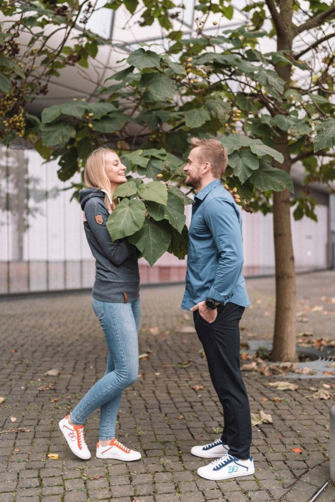 Eine Frau und ein Mann stehen sich auf einem Kopfsteinpflasterweg unter einem Baum gegenüber. Sie trägt einen grauen Hoodie und Jeans, während er das CEDRIQ Blue Shirt von ZAQQ trägt, das gut zu seiner schwarzen Hose passt. Beide tragen stylische Sneakers, lächeln sich herzlich an und versprühen einen mühelosen Charme.