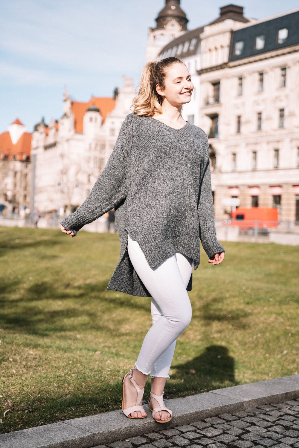 Eine lächelnde Frau steht in einem Park und trägt einen grauen Pullover und eine weiße Hose. Sie ergänzt ihr Outfit mit stylischen CLIQ Nude Sandalen von ZAQQ und genießt den sonnigen Tag im Freien mit historischen Gebäuden im Hintergrund.