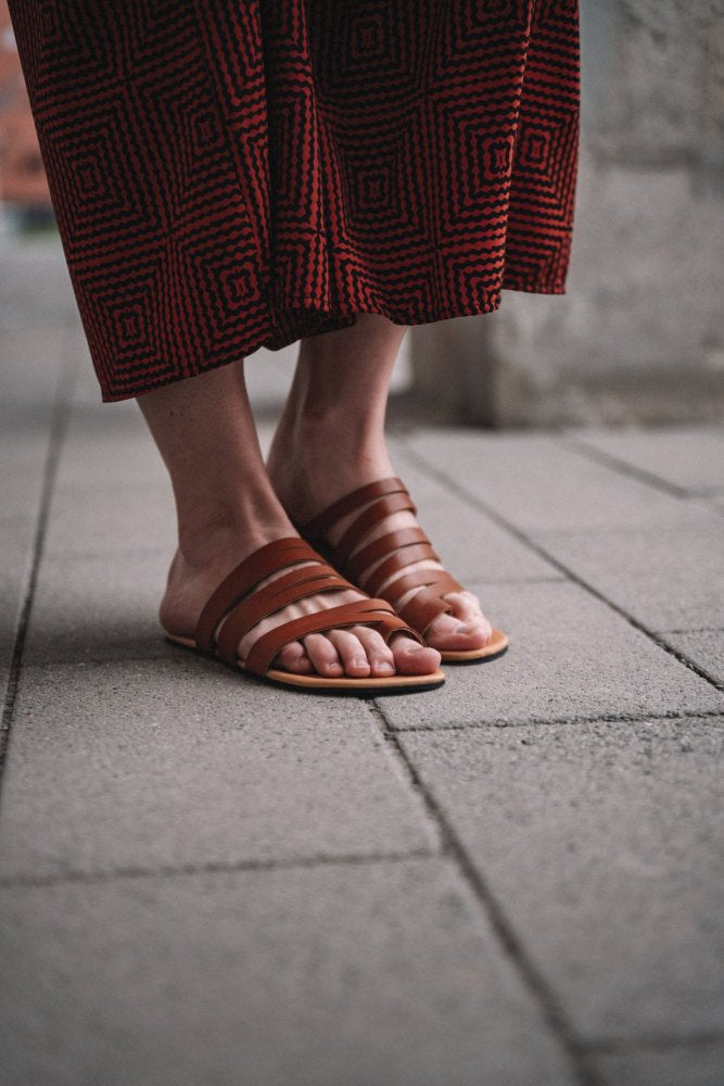 Eine Person, die braune FLAQE-Sandalen von ZAQQ mit rot-schwarz gemusterten Hosen trägt, steht auf einem gefliesten Bürgersteig.