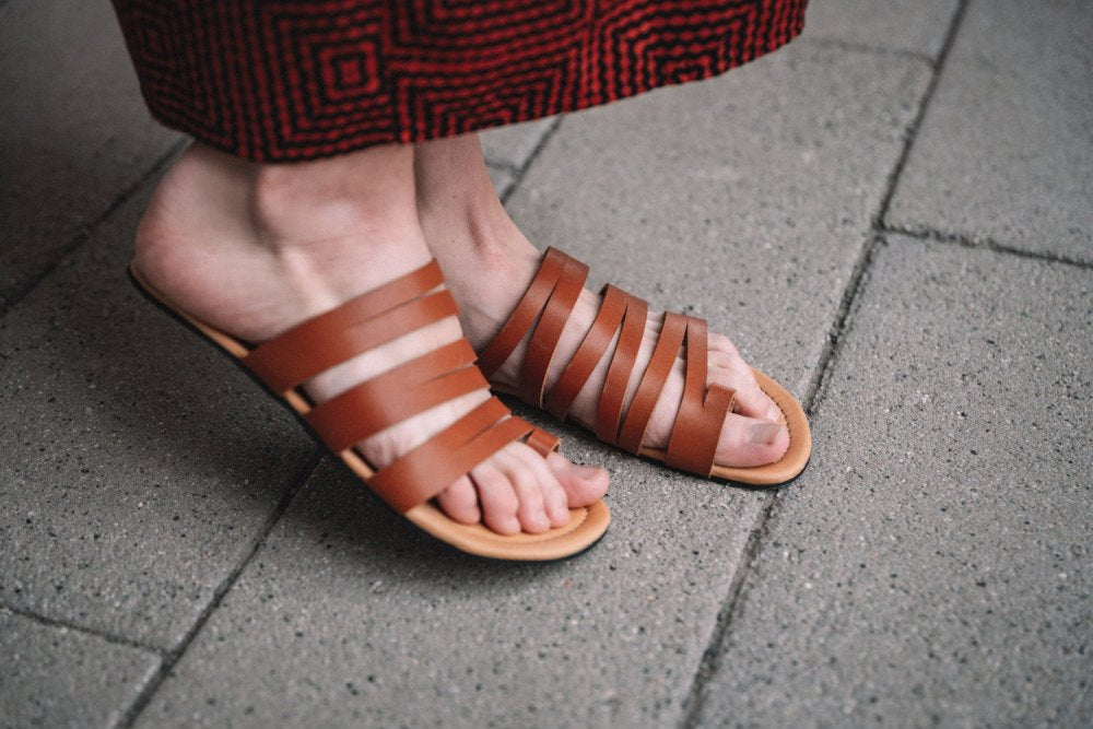 Eine Person, die die FLAQE Brown Sandalen von ZAQQ trägt, steht auf einem Betongehweg. Diese Sandalen aus Nappaleder mit mehreren Riemen ergänzen den rot gemusterten Rock oder das Kleid wunderschön.