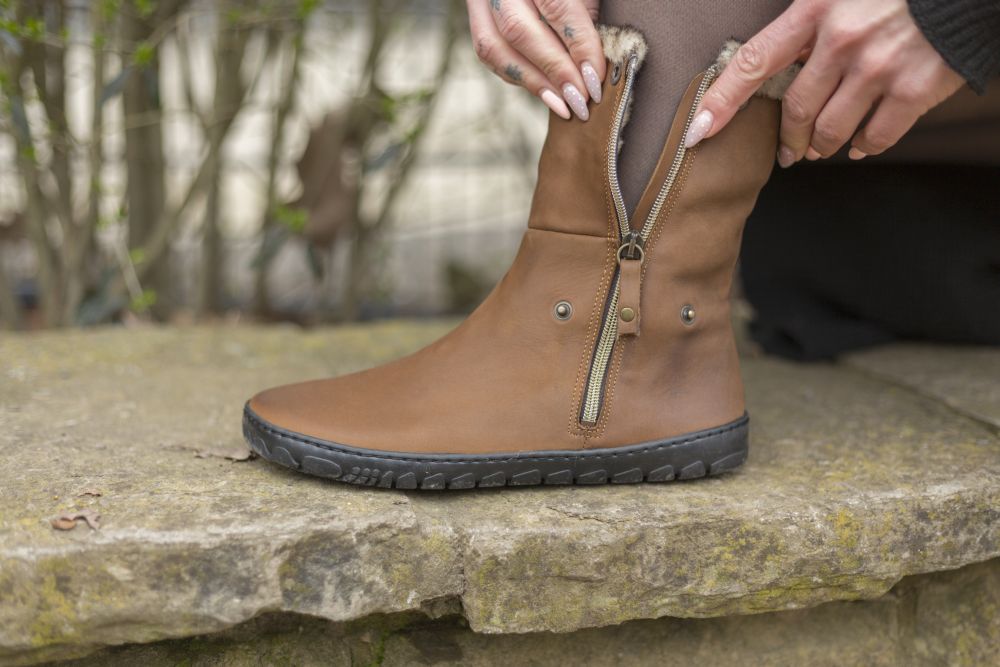 Eine Person sitzt auf einer Steinoberfläche und zieht den Reißverschluss eines HUQ Brown-Stiefels von ZAQQ zu, der ein braunes Velourslederoberteil und eine schwarze Kautschuksohle hat. Der Stiefel hat einen seitlichen Reißverschluss und Knopfdetails. Hände sind sichtbar, die den Reißverschluss zurechtrücken, während das verschwommene Grün im Hintergrund zur ruhigen Atmosphäre beiträgt.