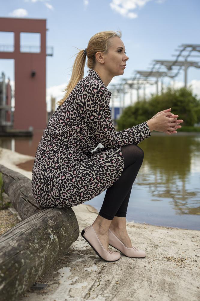 Eine Frau in einem Kleid mit Leopardenmuster und schwarzen Leggings sitzt elegant auf einem Holzbalken neben einem Teich. Ihre blonden Haare sind zurückgebunden und sie trägt bequeme LOOQ Nude Ballerinaschuhe von ZAQQ. Im Hintergrund sind moderne Strukturen und Bäume unter einem teilweise bewölkten Himmel zu sehen.