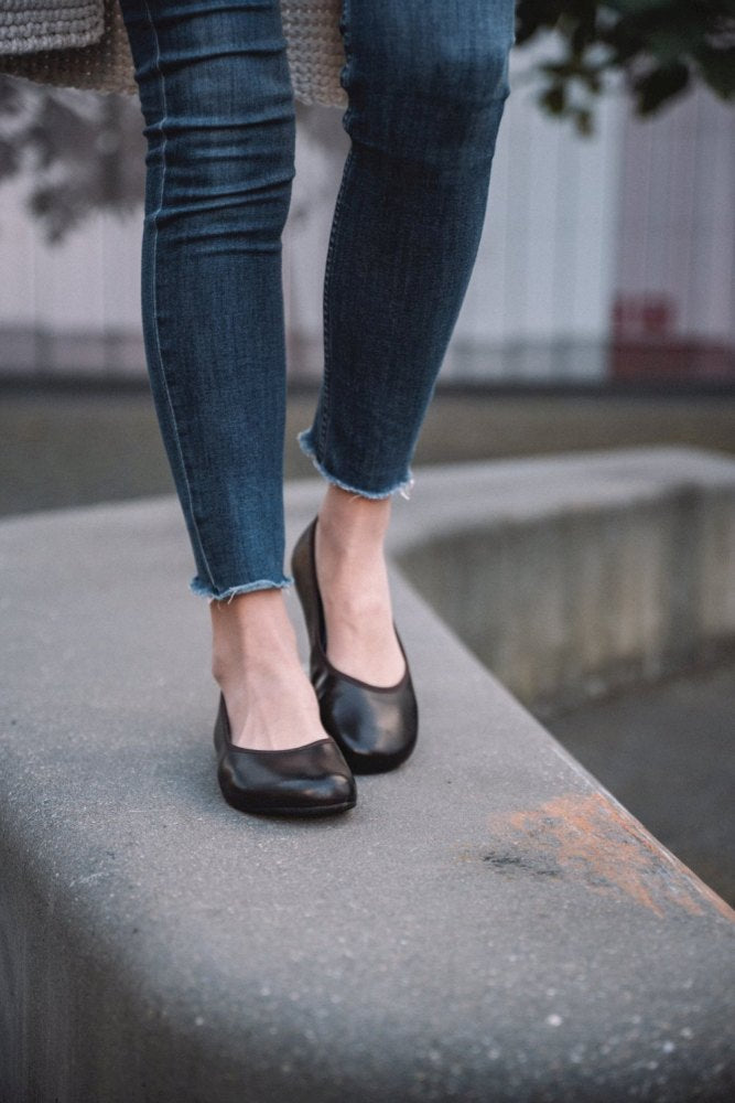 Eine Person in Jeans mit ausgefranstem Saum und dunkelbraunen QARMA-Ballerinas von ZAQQ balanciert auf der Kante einer Betonfläche. Der Hintergrund ist verschwommen und zeigt etwas Grün und einen Teil eines Gebäudes.