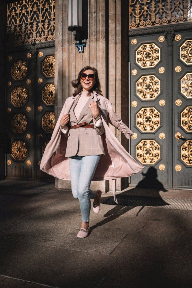 Eine Frau in einem stylischen Outfit posiert glücklich vor verzierten, mit Gold verzierten Türen. Sie trägt einen rosa Mantel, einen karierten Blazer, Jeans und eine Sonnenbrille, dazu QERP Candy-Schuhe von ZAQQ. Das Sonnenlicht wirft einen weichen Schatten auf den Boden.