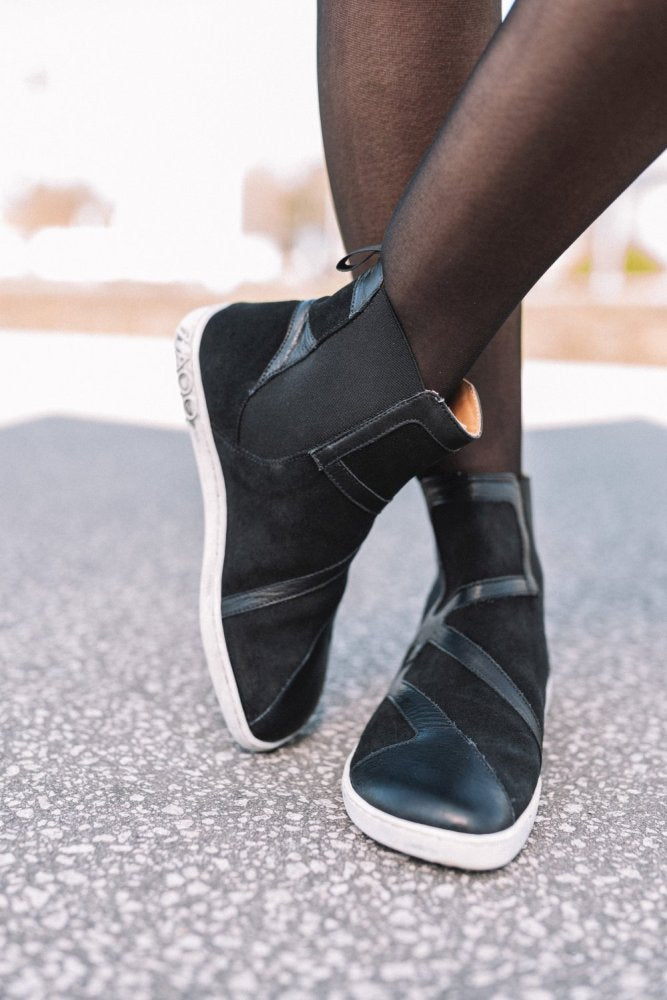 Eine Person trägt die knöchelhohen QEY WEST Black-Sneakers von ZAQQ im Chelsea-Boot-Stil mit weißer Sohle und Riemendesign für zusätzlichen Flair und steht im Freien auf einer strukturierten Oberfläche, während sie schwarze Strumpfhosen trägt. Die Schuhe sind aus Leder und Stoff gefertigt.