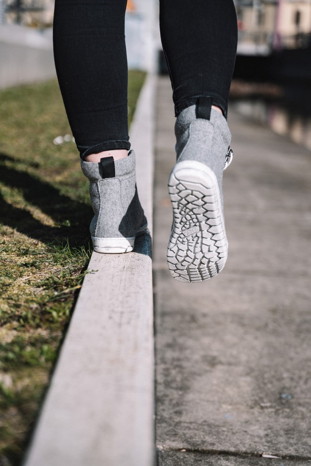 Eine Person trägt QICE Grey High-Top-Sneakers von ZAQQ mit Outdoorsohle und eine schwarze Hose und läuft am Rand eines Gehwegs. Ein Fuß ist vom Boden abgehoben und balanciert vorsichtig. Die Szene spielt sich im Freien ab, auf einer Seite des Weges ist Gras zu sehen.