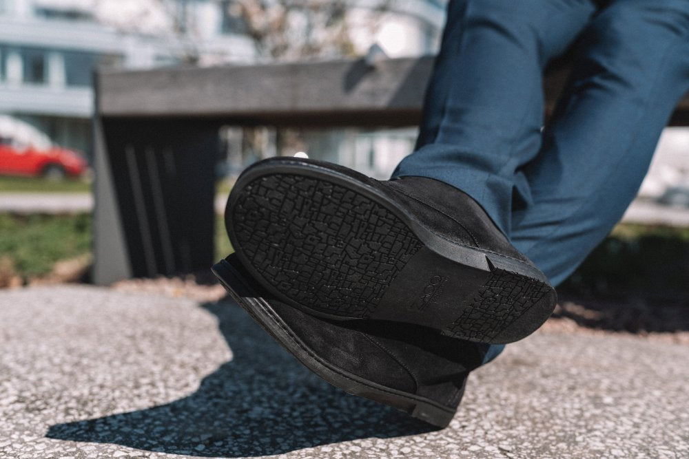 Eine Person in blauer Hose entspannt auf einer Parkbank und präsentiert die Sohlen ihrer schicken schwarzen ZAQQ QLASSY-Schuhe. Im Hintergrund sind ein rotes Auto und Gebäude verschwommen.