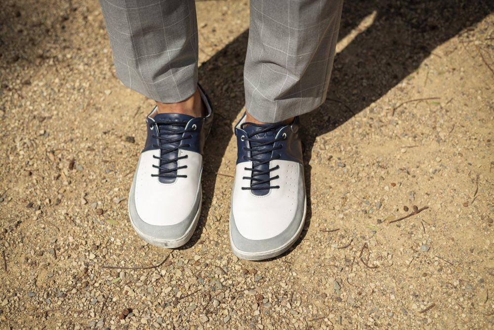 Eine Person, die ZAQQs „QOOL Navy“-Golfschuhe mit grau karierten Hosen trägt, steht auf einem Sandboden und strahlt den Komfort aus, den man von erstklassiger Barfuß-Sneaker-Handwerkskunst erwarten kann.