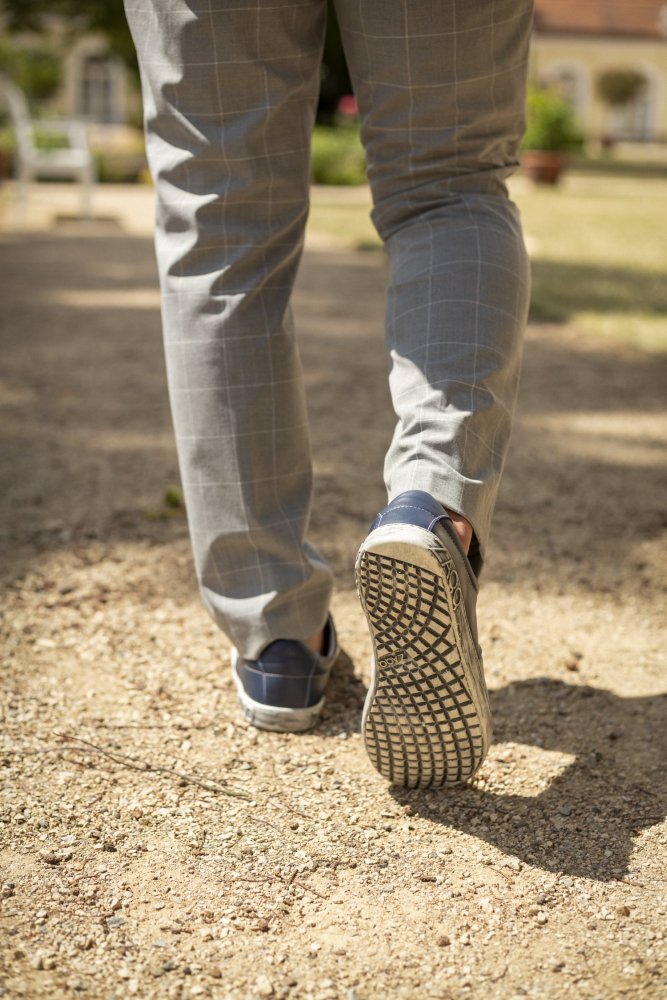 Eine Person in grau karierter Hose und QOOL Navy Sneakers von ZAQQ, bekannt für ihren hervorragenden Tragekomfort, läuft auf einem Kiesweg. Der Fokus liegt auf der unteren Hälfte, wobei die Sohle eines Schuhs sichtbar ist. Im Hintergrund sind unscharfe Bäume und Gebäude zu erkennen.