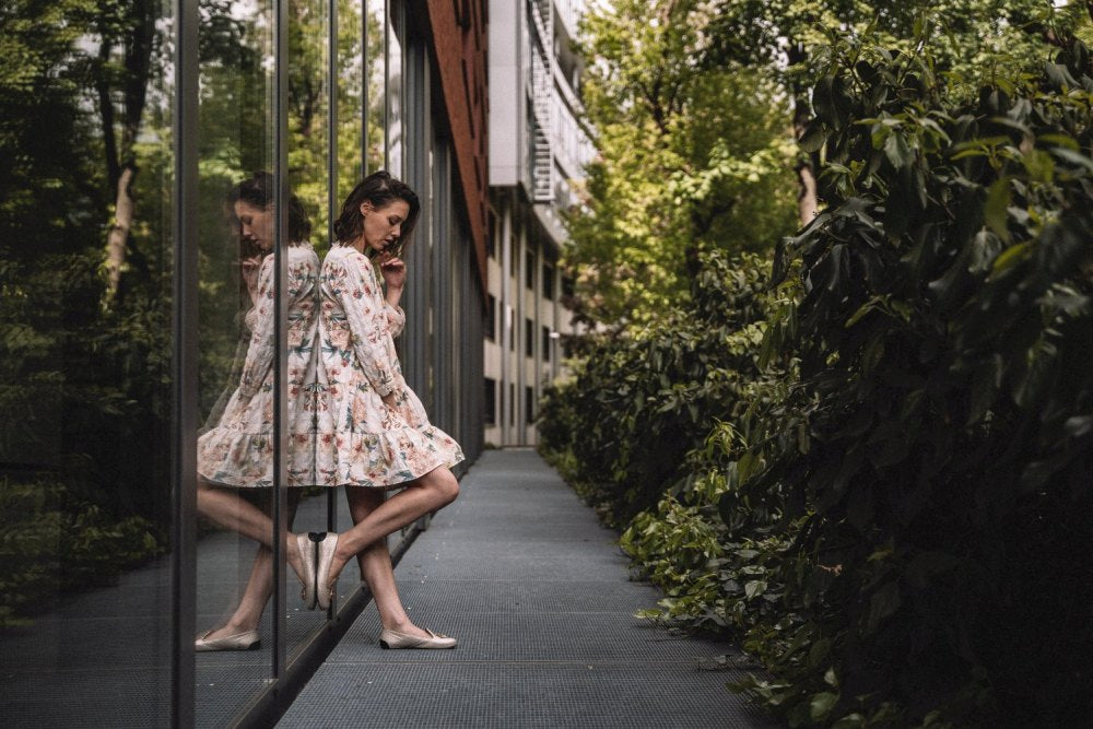 Eine Frau in einem geblümten Kleid lehnt an einem Glasfenster, neben ihr ist ihr Spiegelbild zu sehen. Im Freien, umgeben von üppigem Grün und einem eleganten modernen Gebäude, hält sie ein Glas ZAQQs QOSMO-Champagner in der Hand. Ihr Gesichtsausdruck ist nachdenklich, während sie nach unten blickt.