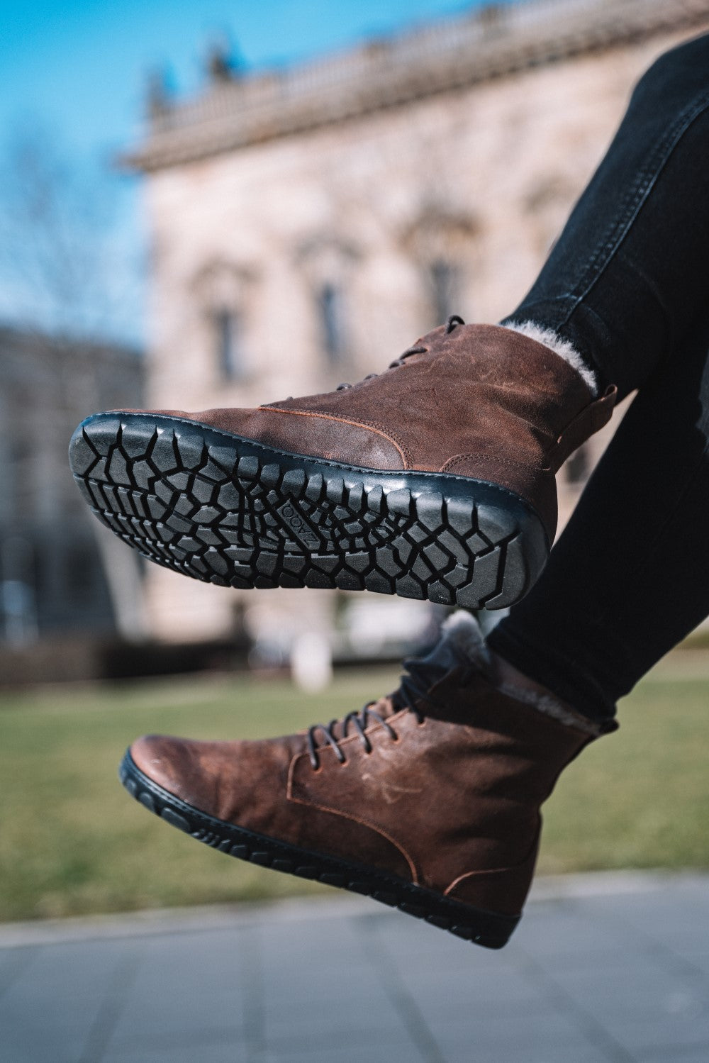 Eine Person trägt QUINTIC Winter Velours Brown Waterproof-Stiefel von ZAQQ mit schwarzen Sohlen und detailliertem Profil und springt mit an den Knien angewinkelten Beinen. Der Hintergrund zeigt ein verschwommenes Steingebäude und grünes Gras unter einem klaren blauen Himmel, wodurch der Fokus auf die Textur der Stiefel gelegt wird.