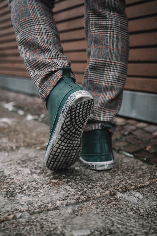 Eine Person trägt die modischen QUITMAN Green Sneakers von ZAQQ aus Velours- und Glattleder und läuft einen Steinweg entlang. Die leuchtend grünen Schuhe und der Boden stehen im Mittelpunkt, grau karierte Hosen tragen zur Ästhetik bei und im Hintergrund ist eine Holzwand zu sehen.