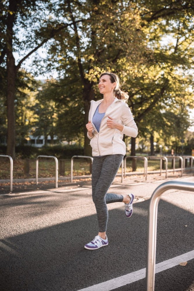 Eine Person joggt auf einem gepflasterten Weg in einem Park, umgeben von Bäumen mit grünem Laub. Sie trägt einen weißen Kapuzenpullover, graue Leggings und die RIQ Purple-Sneaker von ZAQQ mit Akzenten aus Veloursleder und atmungsaktivem Meshgewebe und genießt die Bewegung im Freien an einem sonnigen Tag.