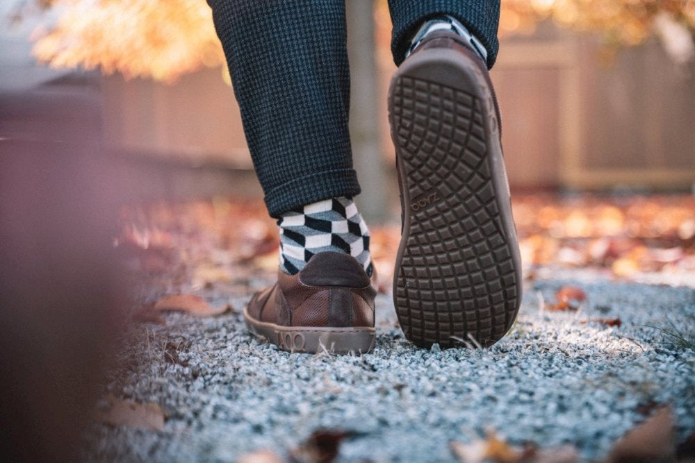 Eine Person, die auf einem Kiesweg mit gefallenen Herbstblättern spazieren geht, betont den Komfort ihrer ZAQQ RIQO Brown Barfußschuhe, während sie schwarz-weiße Socken mit geometrischem Muster trägt. Die Szene spielt vor einem Hintergrund mit einem Holzzaun und verschwommenen Bäumen, die mit leuchtend orangefarbenen Blättern geschmückt sind.