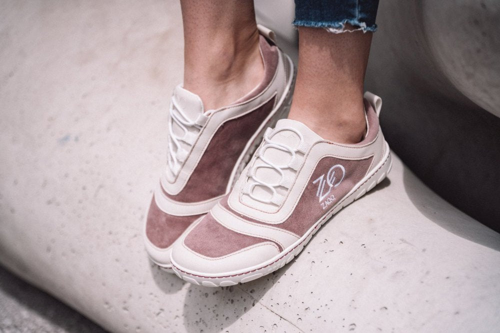 Nahaufnahme einer Person, die stylische ZAQQ SQILL Rose Sneaker mit rosa Wildlederakzenten und weißen Schnürsenkeln trägt. Diese Schuhe sind aus veganer Mikrofaser gefertigt und haben ein aufgesticktes Logo an der Seite. Die Person steht auf einer strukturierten Steinoberfläche, der Saum ihrer Jeans ist leicht ausgefranst.