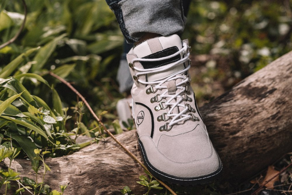 Eine Nahaufnahme einer Person, die den TERRAQ Beige Waterproof Wanderschuh von ZAQQ trägt und in einer üppigen, grünen Waldlandschaft auf einen umgestürzten Baumstamm tritt. Dieser Outdoor-Schuh verfügt über Metallösen und weiße Schnürsenkel und verbindet auf schöne Weise Funktionalität mit Stil.