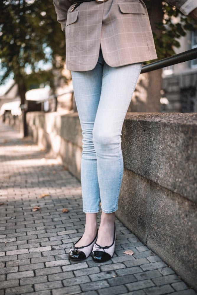 Eine Person in hellblauer Jeans und ZAQQs TWIST Varnish Nude Flats mit bequemer Kautschuksohle steht auf einem Kopfsteinpflasterweg. Sie lehnt an einer Steinmauer, im Hintergrund eingerahmt von Bäumen und Gebäuden, und kombiniert ihr Outfit mit einem karierten Blazer.