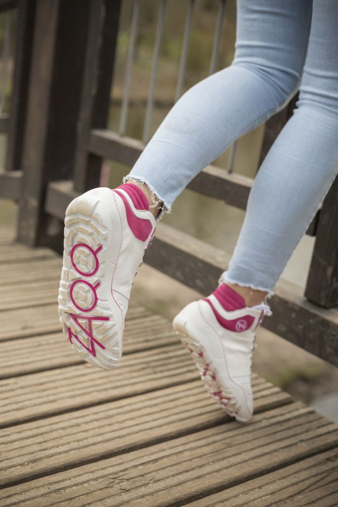 Eine Person, die ZAQQs VAQATION Pink Waterproof-Sneaker trägt, die das KOOL-Design auf der Sohle tragen, sitzt in hellblauen Jeans auf einem Holzgeländer. Der Hintergrund zeigt verschwommenes Grün und Wasser und verbindet Komfort und Stil perfekt, wie die Barfuß-Leichtigkeit der Barfußschuh-Designs.