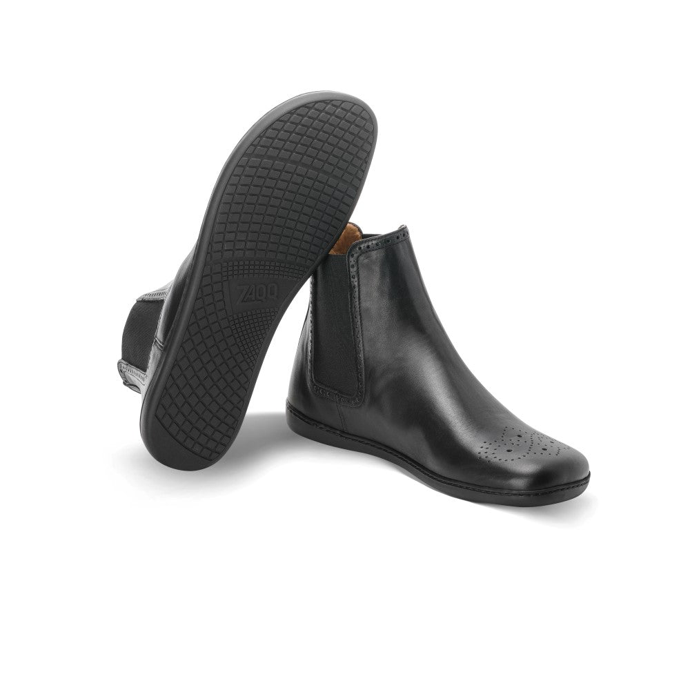 Ein Paar ZAQQ EQUITY Brogue Stiefeletten aus schwarzem Leder mit elastischen Seiten. Ein Chelsea-Boot steht aufrecht und hebt die perforierten Details an der Spitze hervor, während der andere Boot geneigt ist, um das komplizierte Profilmuster auf seiner Sohle zu zeigen.