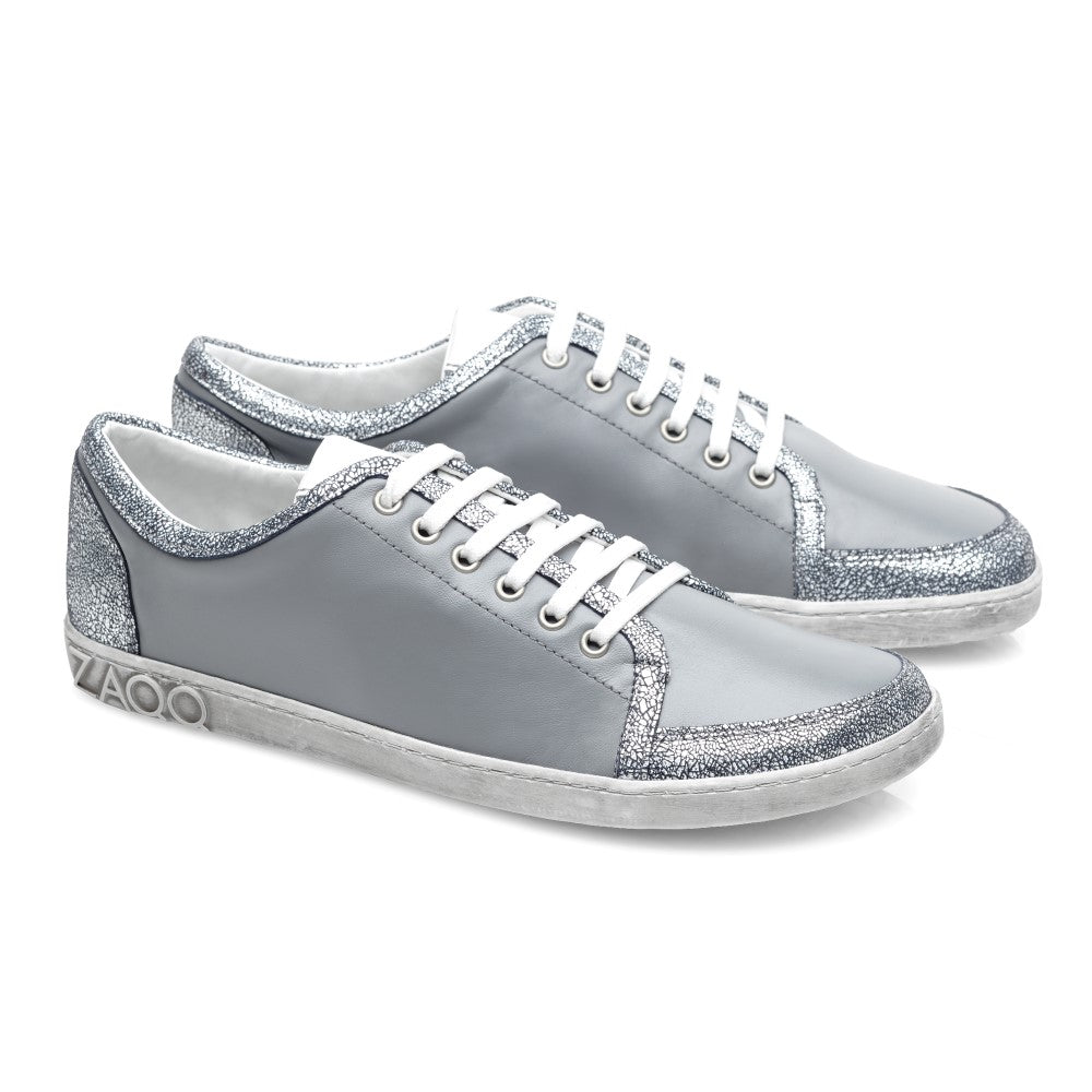 Die TIQQ Grey Silver Sneaker von ZAQQ bestechen durch ihr schlichtes Design aus weichem Nappaleder mit weißen Schnürsenkeln und silbernen Glitzerakzenten, ergänzt durch weiße Sohlen und ein dezentes Logo an der Ferse, und bieten die perfekte Mischung aus Stil und Komfort für jeden Anlass.